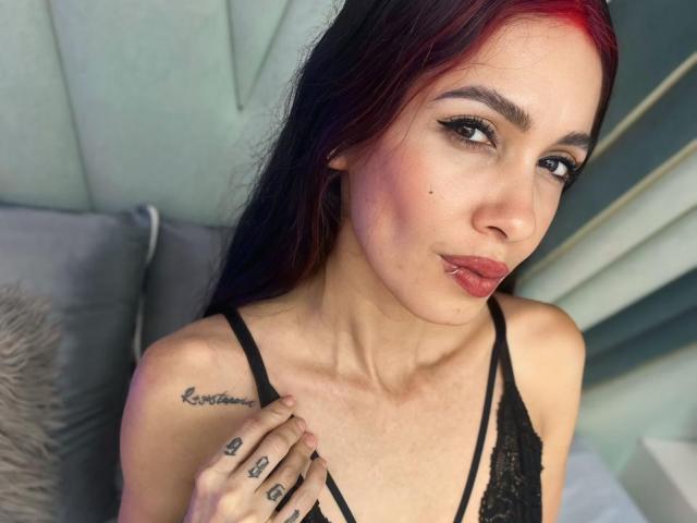 KathySteven, con i capelli tinti di rosso, una maglia di pizzo nero e un piercing al labbro, mostra un tatuaggio sulla spalla sinistra e dita con anelli, con un'espressione accigliata e un trucco leggero, in un ambiente che sembra una camera da letto..