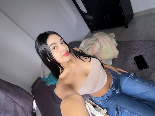 TinaBurnny with long black hair, tan strapless top, and blue jeans, sitting on bed, showing her tits and holding phone, blanket and clothes in background, red lipstick.