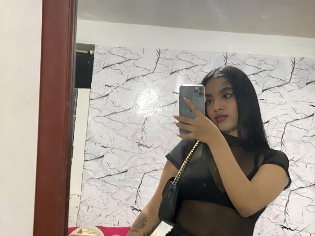TinaBurnny with long black hair, wearing a sheer black top, holding a phone in front of a mirror, showing her tits and tattoo on her right arm.