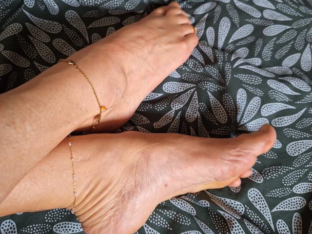FrenchieLilieFleur's feet with gold anklets, showing slight wrinkles and tan skin, resting on a dark green patterned surface. Her toes are slightly curled and relaxed.