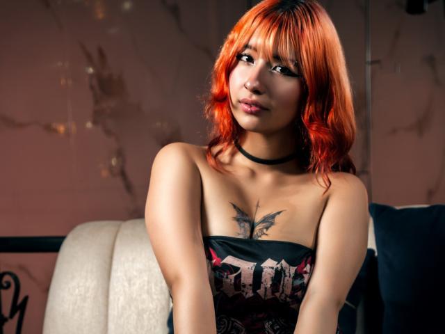 Emily Lenox, com cabelo laranja, colar preto e tatuagem acima dos seios, sentada em um sofá branco, olhando para a câmera, plano médio, iluminação suave..