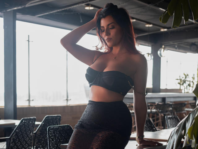 ZoeeMar, avec ses cheveux foncés, portant un soutien-gorge noir sans bretelles et une jupe noire texturée, appuyée sur une table, la main dans les cheveux, mettant en valeur ses seins et ses fesses dans une pièce baignée de soleil..
