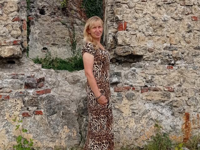 TrinaAdair stands against a rough stone wall, wearing a brown and white leopard-print dress, with blonde hair and a slight smile, holding her hands in front.