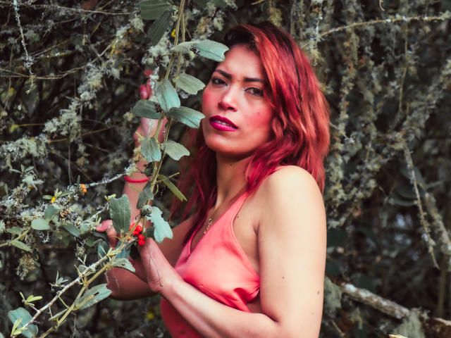VioletaSexiest with red hair, pink tank top, holding a plant, looking at camera, pink lipstick, slight blush, forest background, red berries in her hand, right arm raised.