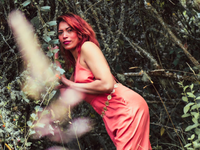 VioletaSexiest with red hair, wearing a pink dress, bending forward in a forest, holding branches, showing her ass, with a sexy, confident smile, and vibrant red lipstick.