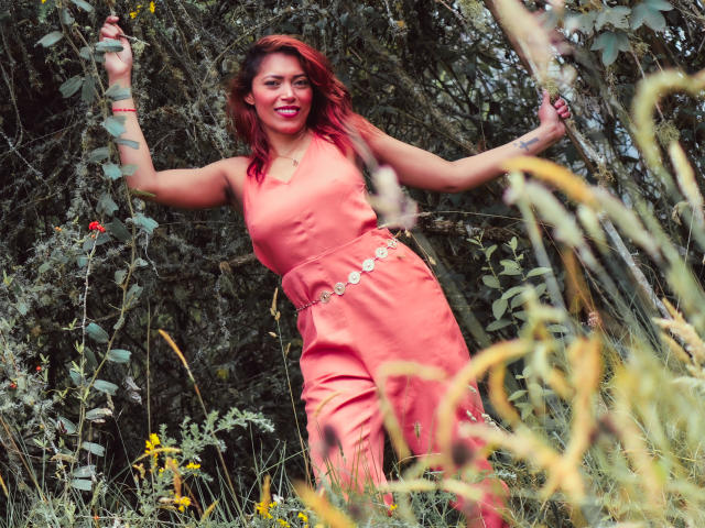 VioletaSexiest with red-tinted hair, wearing an orange dress with a gold belt, standing in a wild grassy area, smiling, holding a branch, arms raised, showing tattoos.