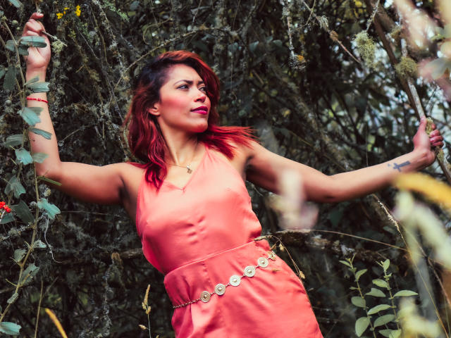 VioletaSexiest with red hair, wearing a pink dress and a gold belt, stands in a bushy, green outdoor setting, arms raised, looking to the side with a confident expression.