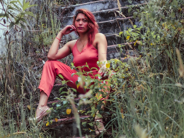 VioletaSexiest with red hair, wearing a red dress, sitting on old, rusty stairs, surrounded by green plants, one hand on her chin, looking thoughtful, with visible tits.