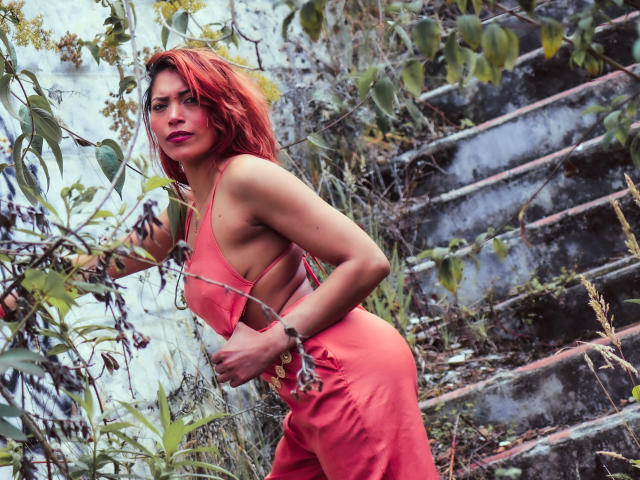 VioletaSexiest with red hair, wearing a pink dress, leaning against a leafy branch, showing her sideboob, and a cute ass, in a rustic outdoor setting.