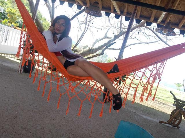 AlisonCold with long dark hair, white long-sleeve shirt, black shorts, and black sandals, sitting in an orange hammock, smiling, showing her ass, under a wooden roof.