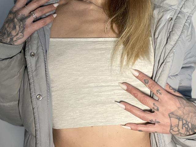 DaneeSoHot's small tits in a white crop top, wearing a grey jacket, hands with tattoos, long blonde hair, and long nails, standing with a confident pose.