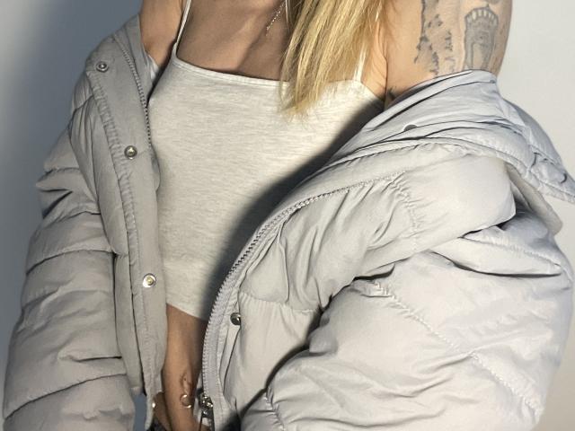 DaneeSoHot's grijze, opvallende jas is van haar schouders gevallen, waardoor een witte top en tatoeages op haar rechterarm zichtbaar zijn. Blond haar, een zichtbare ketting en een lichte bruine huid..