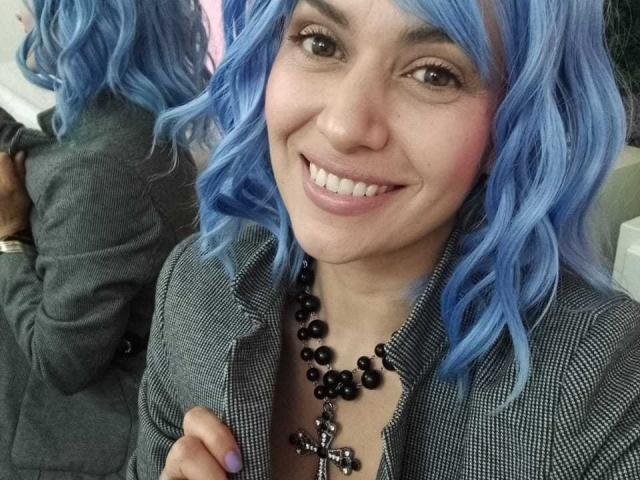 LilaaSalmon with blue curly hair, smiling, wearing a black beaded necklace with a cross, and a checkered gray blazer. Boobs not visible, ass not visible.
