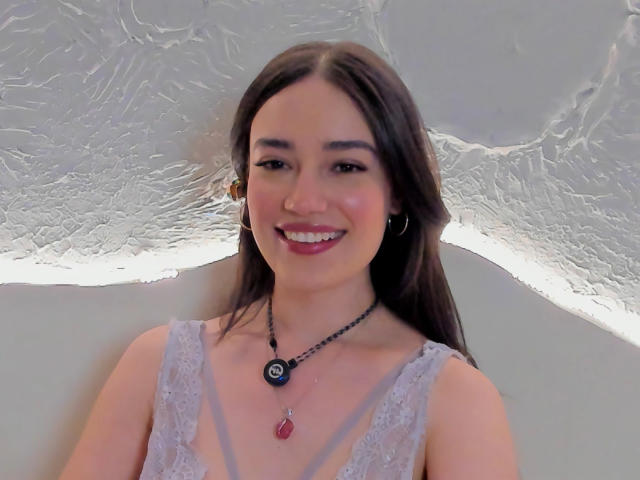 MariaLoft tem cabelos castanhos longos, um sorriso adorável, e está vestindo uma blusa de renda com um colar que tem um pingente preto e uma gema vermelha. Ela está sorrindo para a câmera..