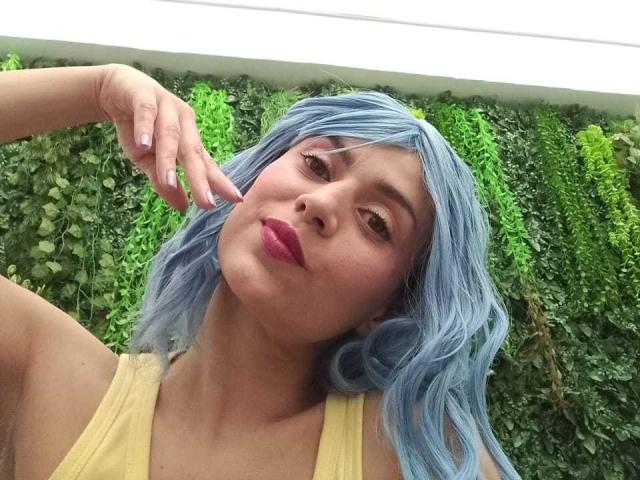 LilaaSalmon with blue hair, pouty pink lips, and a yellow tank top, touching her cheek, has a cute face and a slightly upturned nose, looking at the camera.
