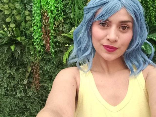 LilaaSalmon with blue hair, wearing a yellow tank top, pink lipstick, and brown eyes, taking a selfie in front of a green leafy wall. Boobs visible, looking cute.