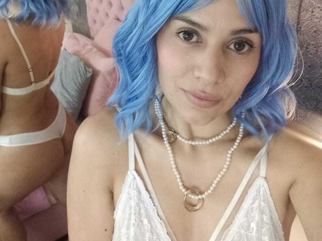 LilaaSalmon with blue hair, white lace top, pearl necklace, and white bra, showing her back and ass in a mirror, looking at the camera with a slight smile.