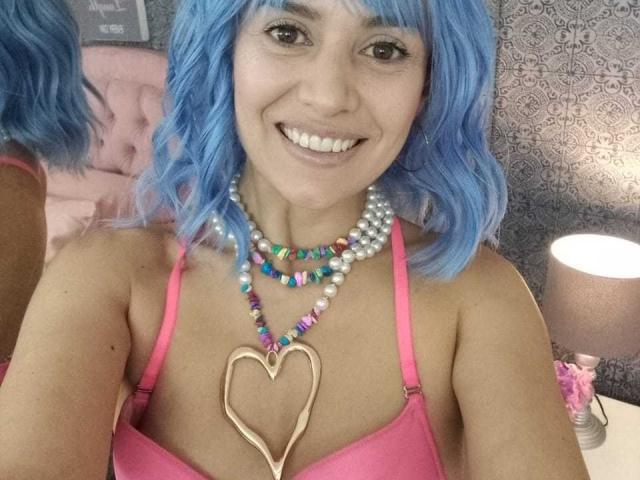 LilaaSalmon with blue curly hair, pink bra, multiple bead necklaces, heart-shaped pendant, smiling, showing teeth, brown eyes, fair skin, cute, with a pink lamp on the right.