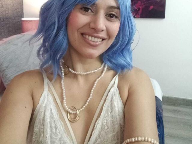 LilaaSalmon with blue wavy hair, smiling, wearing a lace white top, pearl necklace, and bracelet, showing her tits and cute face.