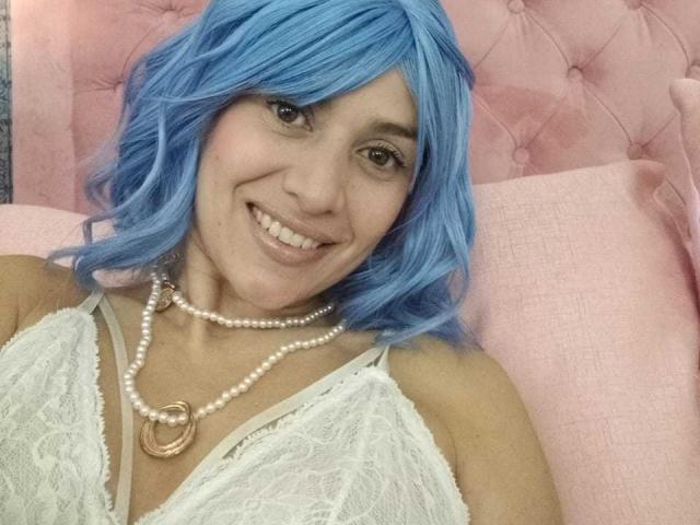 LilaaSalmon with blue hair, smiling, wearing white lace top, pearl necklace, and gold ring, showing her tits. Pink tufted background.