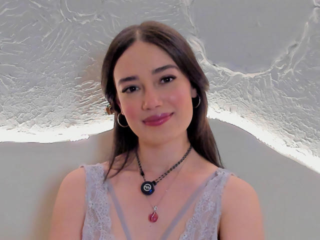 Maria, con lunghi capelli castani, indossa una maglia con maniche in pizzo, orecchini a cerchio, una collana nera con un ciondolo circolare e rossetto rosa, sorridendo alla fotocamera..