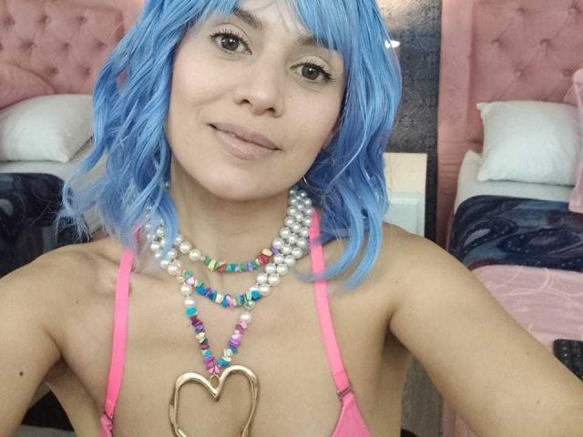 LilaaSalmon with blue wig, pink bra, pearl and rainbow bead necklace, heart pendant, smiling, brown eyes, light skin, small tits, bed with pink headboard, white pillow, dark blanket.