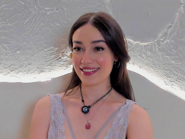 MariaLoft, avec ses longs cheveux bruns, souriante, portant un haut en dentelle, un collier noir avec un pendentif en forme de goutte rouge, et de petites boucles d'oreilles, a de jolies seins et un joli nez..
