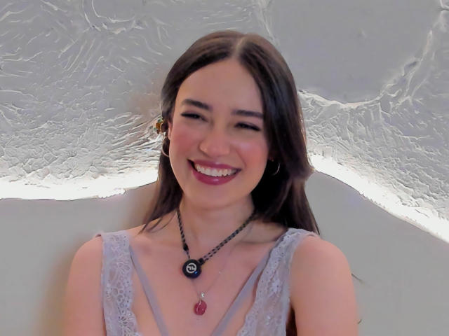 MariaLoft, con i capelli castani, sorridente, indossa una camicetta in pizzo, una collana nera con un ciondolo rosso e orecchini, ha un bel seno, una pelle chiara e rossetto rosa..