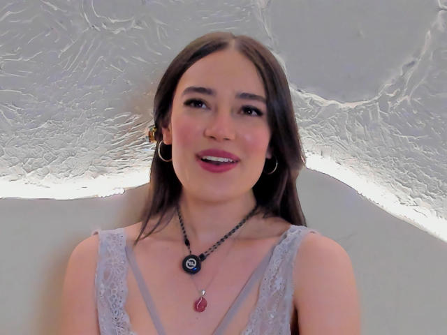 MariaLoft has long brown hair, wearing a gray lace dress, hoop earrings, black necklace with a red gem, smiling with pink lipstick, and light skin.