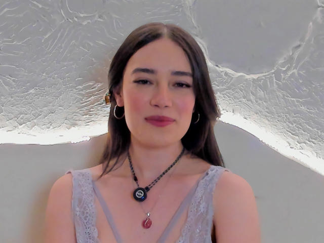 Maria, con cabello largo y castaño, vestida con un vestido gris de encaje, aretes grandes, un collar negro con colgantes azules y rojos, sonriendo a la cámara..