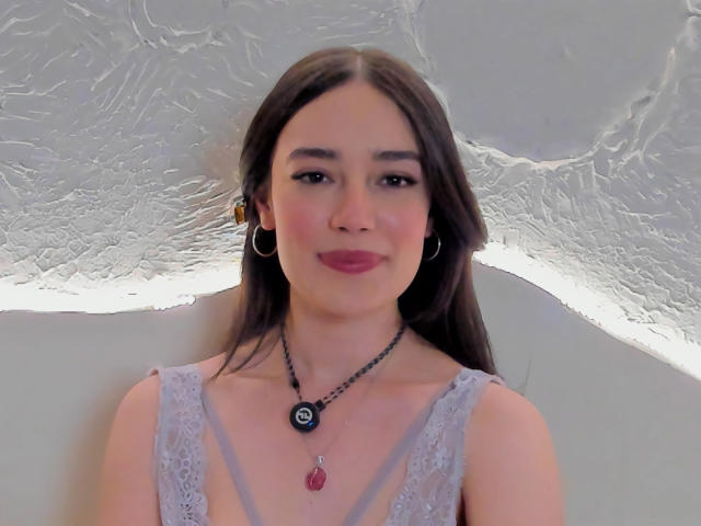 Maria, con lunghi capelli castani, indossa un abito grigio in pizzo, orecchini a cerchio, una collana nera con un ciondolo blu e una pietra rossa, e rossetto rosa, sorridendo alla fotocamera..