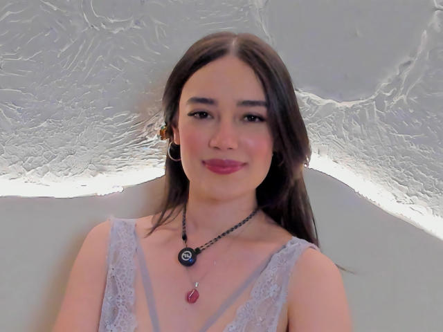 MariaLoft heeft lang bruin haar, roze lippenstift, oorringen, een zwarte ketting met een rode steen, en draagt een top van kant zonder mouwen, terwijl ze glimlachend naar de camera kijkt..