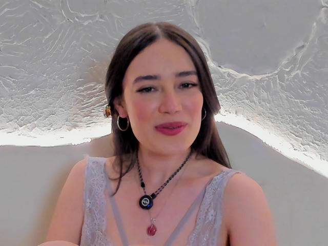 MariaLoft a de longs cheveux bruns, elle porte un haut gris en dentelle, des boucles d'oreilles cerceaux, un collier noir avec un bijou rouge, et du rouge à lèvres rose, elle sourit doucement avec un visage mignon..