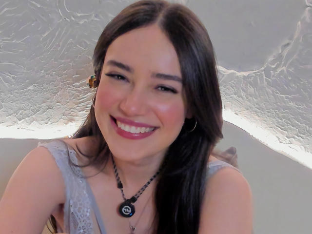 MariaLoft ha lunghi capelli castano scuro, rossetto rosa e un grande sorriso che mostra denti bianchi. Indossa una maglia grigia in pizzo e una collana nera con un ciondolo circolare..