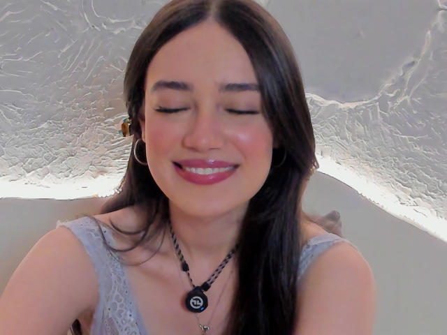 MariaLoft, les yeux fermés, souriante, avec un rouge à lèvres rose, des cheveux bruns, des boucles d'oreilles cerceaux, un haut en dentelle gris, un collier noir, un plafond blanc texturé et un petit pendentif noir et blanc..