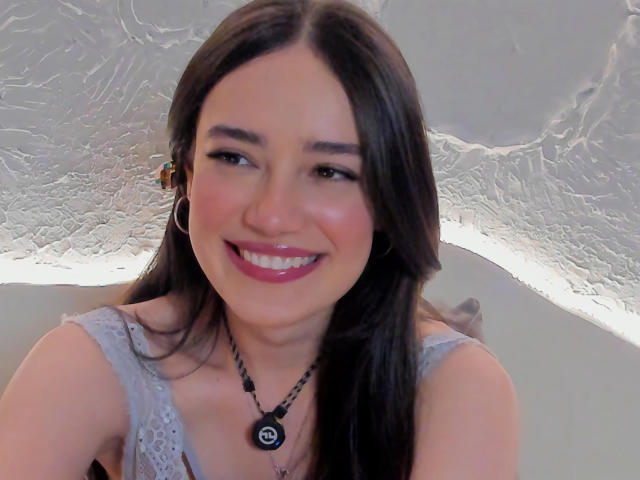 MariaLoft, sorrindo com batom rosa, cabelo castanho, brincos de argola, colar preto, blusa de renda, mostrando os seios levemente, com um teto branco texturizado ao fundo..