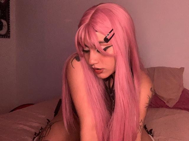 DiynaStar with long pink hair, tattoos, and black hair clips, kneeling on a bed, looking down, showing off her small tits and ass, with soft pink lighting.