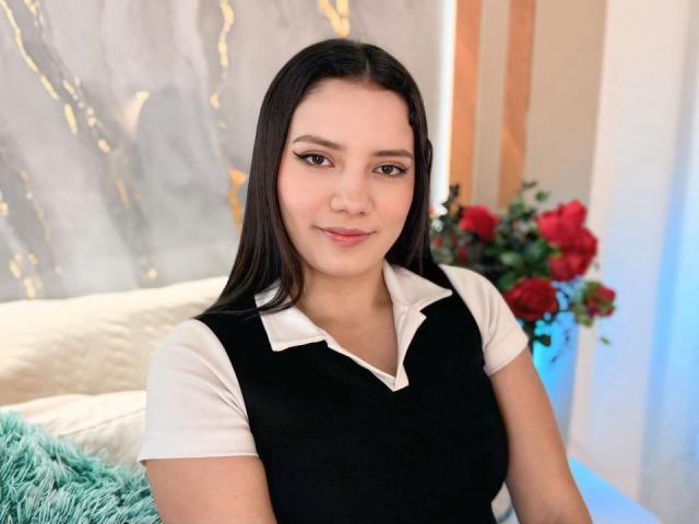 JulietteBlossom with long black hair, wearing a black and white polo shirt, has small tits, light skin, and a slight smile, sitting in a brightly lit room.