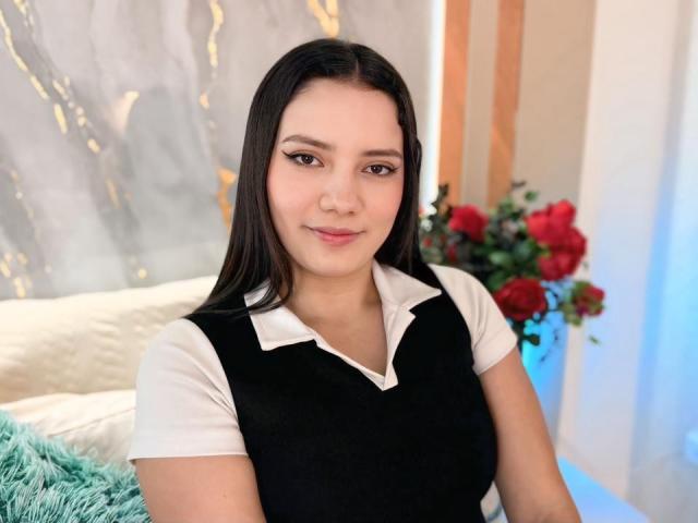 JulietteBlossom with long black hair, wearing a black sleeveless top over a white shirt, smiling softly, having cute brown eyes and a small nose, sitting near red flowers.
