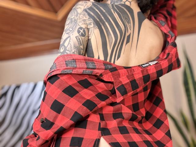 HannaBorbon's back, tattooed with black tribal patterns, in a red and black checkered shirt, off-shoulder, exposing her bare shoulder and part of her upper back.