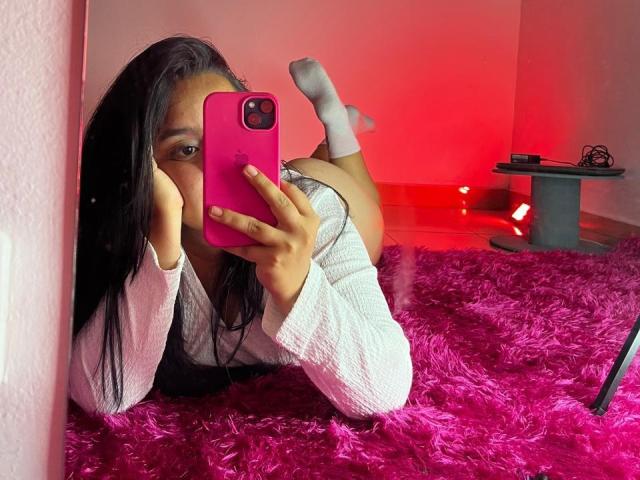 SammyTitss lying on a pink shaggy rug, taking a selfie with a pink phone, wearing a white long-sleeve shirt, and white socks, showing her ass.