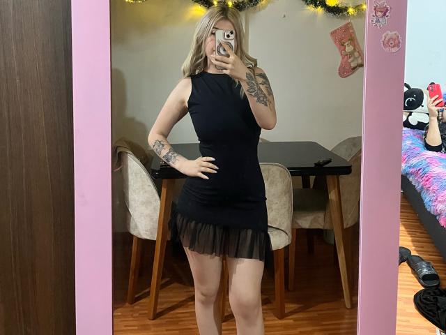 SammyViolet, vestida con un vestido negro, sin mangas y con volantes, mostrando tatuajes en su brazo y mano, se toma un selfie con su teléfono, con su trasero ligeramente visible, y tiene el cabello rubio..