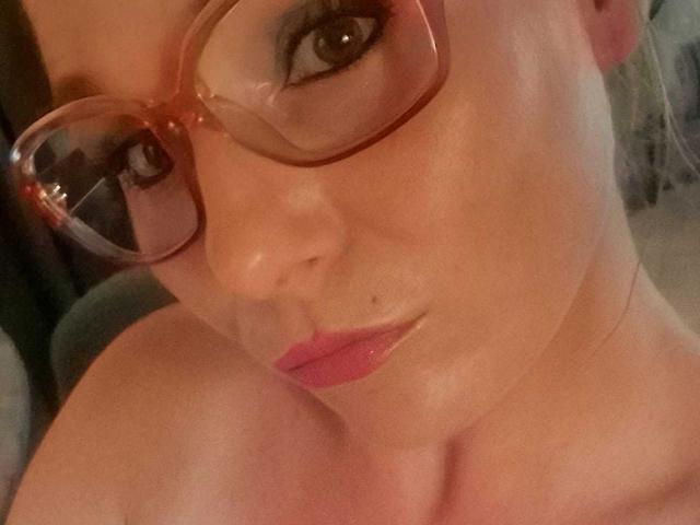 DevilsAngel met glanzende huid, roze lippen en een bril, die haar blote schouders laat zien en met een subtiele glimlach naar de camera kijkt. Borsten zichtbaar, geen andere lichaamsdelen..