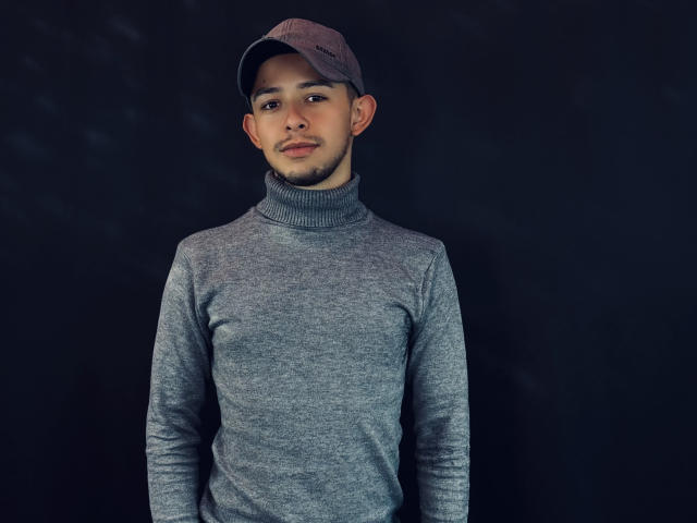 AlecAndrews with a light brown cap, short beard, and medium skin tone, wearing a grey turtleneck sweater, standing against a dark background, looking slightly to the left.