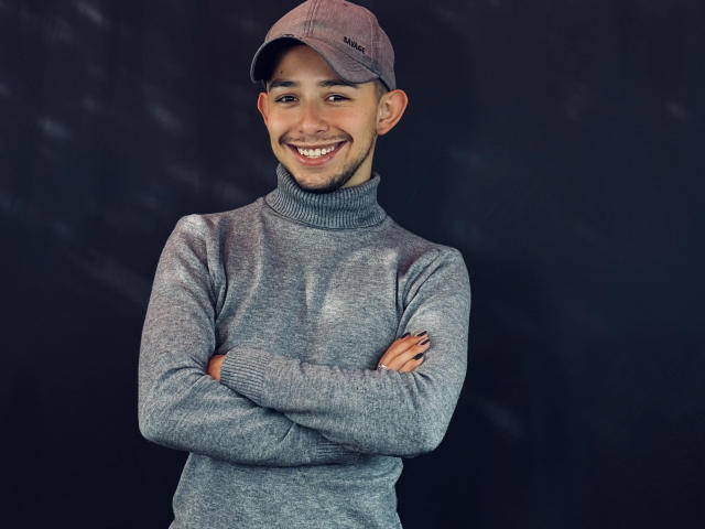 AlecAndrews, with a gray turtleneck and matching cap, stands smiling with arms crossed, showing a cute face and short dark hair, against a dark background.