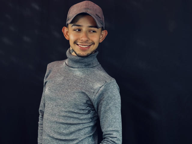 AlecAndrews with a cute smile, wearing a gray turtleneck and pinkish-brown cap, has a medium build, light brown skin, and short dark hair, standing against a dark background.