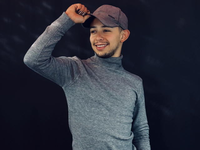 AlecAndrews with a cute smile, wearing a gray turtleneck and matching cap, touching his cap with a relaxed hand, standing against a dark background.
