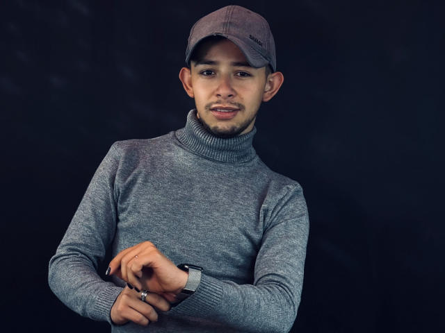 AlecAndrews with a grey turtleneck, grey cap, and dark background, crossing his arms, showing a ring, and wearing a watch, looking at the camera with a slight smile.