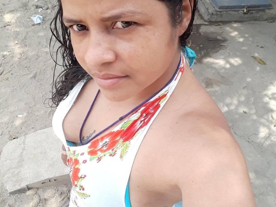 FabiolaX with medium brown skin, wet black hair, wearing a white halter top with red floral patterns, showing her bare shoulders and a purple necklace, looking up at the camera.