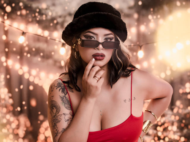 MariGonz, avec ses cheveux noirs et ondulés, portant un chapeau en fourrure noire, un top rouge en forme de spaghetti et des lunettes, touchant ses lèvres avec un doigt, montrant des tatouages et des piercings, dans une pose sexy..